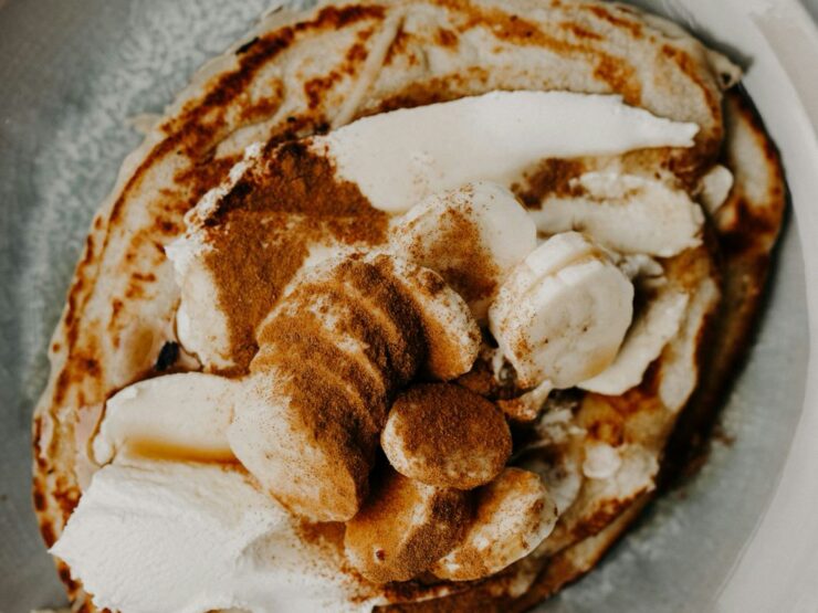 A pancake on a pale plate topped with banana slices, spoonfuls of cream and a heavy dusting of cinnamon