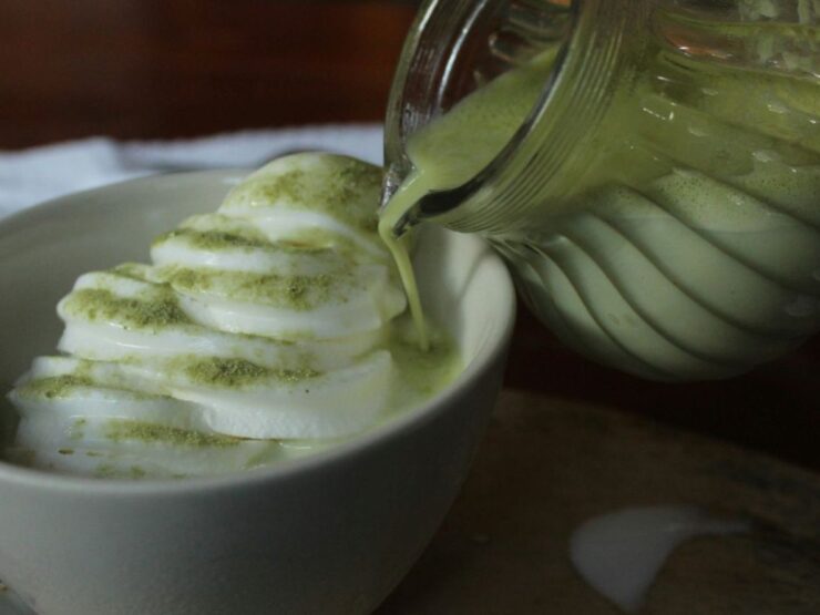 Herbed green yogurt dressing being poured from a glass jug over overlapping cucumber slices in a white bowl