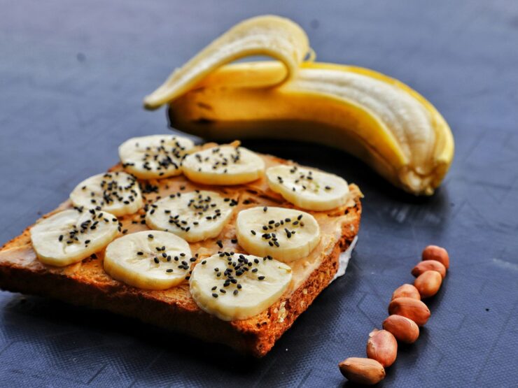 A slice of toast spread with peanut butter, topped with banana rounds and black sesame seeds, a peeled banana and loose peanuts alongside