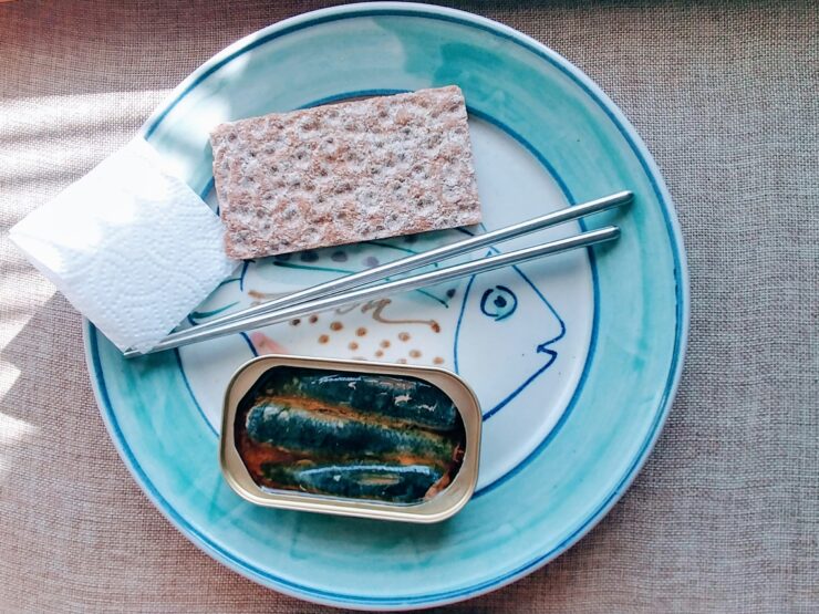 An open tin of sardines, a rye crispbread and a fork on a blue-rimmed plate painted with a fish